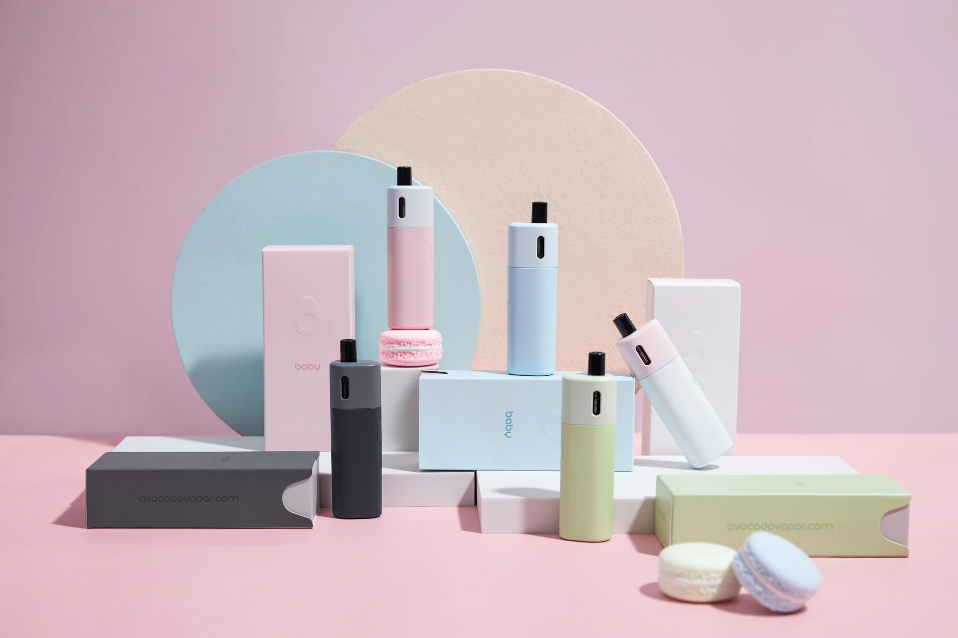 Still-life photography of pastel-finish vaping devices, built on coloured paper geometry with fruit and floral props.